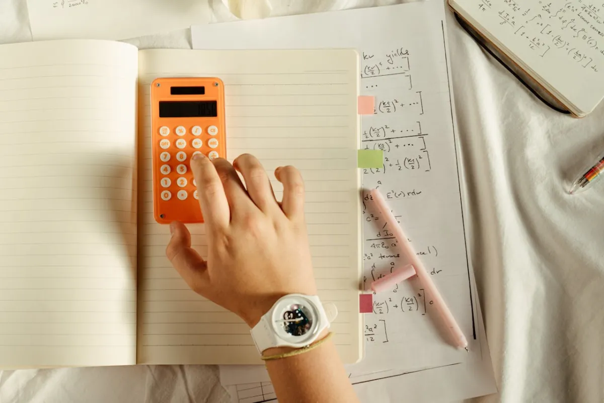 Student working through mathematics calculations with notebook and calculator on desk